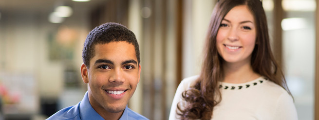 young man and woman stand in their new workplace smiling proudly to camera .